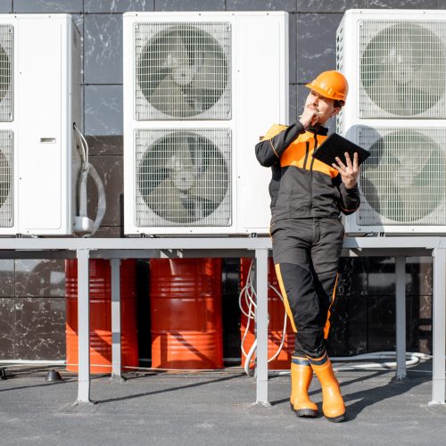 Egineer in protective workwear designing with digital tablet, while standing near the outdoor units of the air conditioner or heat pump